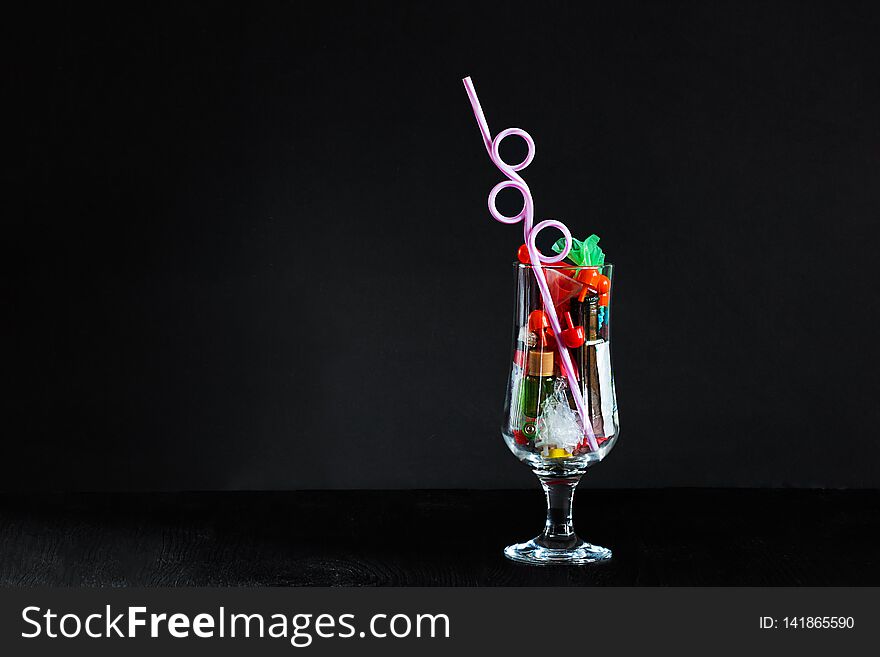 Coctail glass with plastic straw full of plastic and battery waste as imitation of real drink on horozontal black background with copy space. Ecology concept of getting pollution to drinks and food