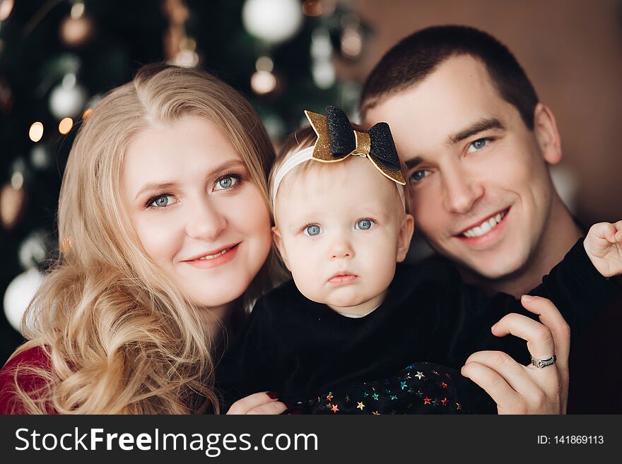 Portrait of happy family smiling at camera against unfocused illumination of decorated christmas tree. Mother, father and baby girl with bow headband. Portrait of happy family smiling at camera against unfocused illumination of decorated christmas tree. Mother, father and baby girl with bow headband.