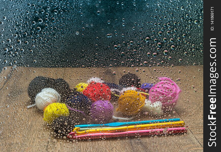 Leftover yarn on a wooden table with crochet hooks, seen through a window with rain drops