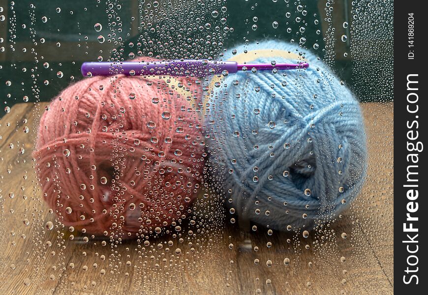 Pink and blue wool with a purple crochet hook on a wooden surface. The window has raindrops on it. Room for text. Pink and blue wool with a purple crochet hook on a wooden surface. The window has raindrops on it. Room for text.