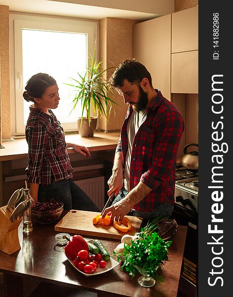 Beautiful young couple in kitchen at home while cooking healthy food. Man cuts a pepper. Woman looks at her husband. Scene from