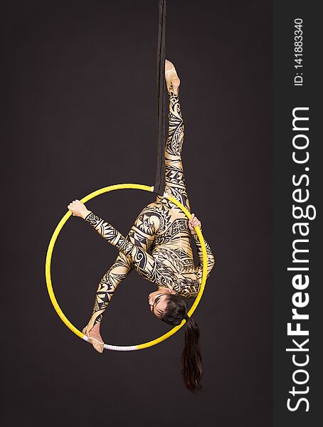 A young girl in a suit with a print performs acrobatic elements in the air ring. Studio shooting performances on a black background