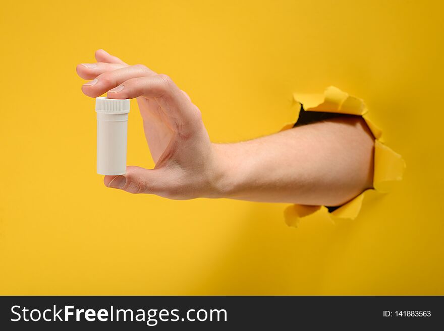 Hand holding a white bottle of pills through a hole torn in yellow paper wall. Innovative medicine or supplement.