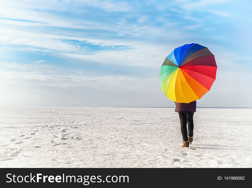 Woman with rainbow colored umbrella walking on frozen sea. Bright colored winter landscape