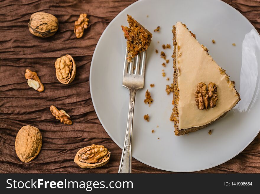 Top view of homemade carrot cake with cream cheese topping decorated with piece of walnut placed on white plate with silver fork on brown tablecloth with walnuts and nutshell next to the plate