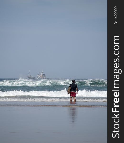A young man stands on the coastline checking surf conditions, a ship can be sought on the background
