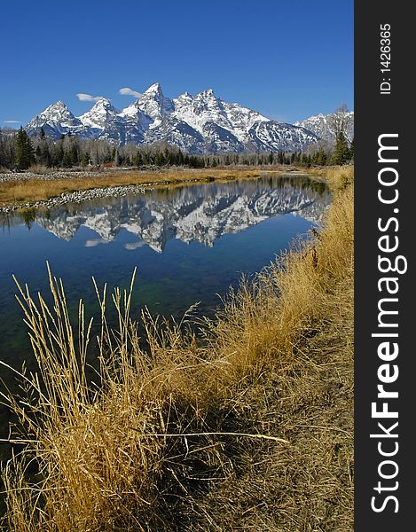 Teton mountain range reflecting in river water with surrounding rocks and plants