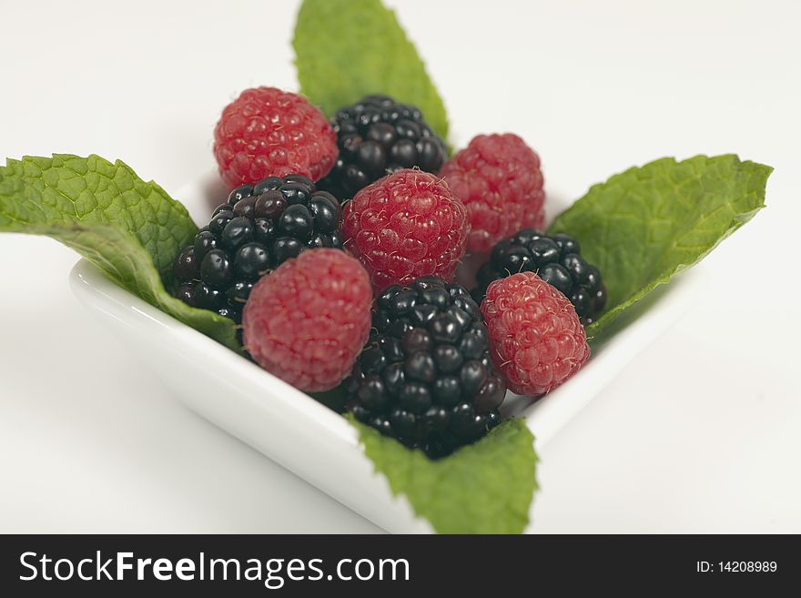 Fresh raspberries and blackberries in a square dessert dish garnished with mint leaves on a white background.