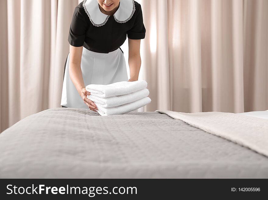 Young maid putting stack of fresh towels on bed in hotel room