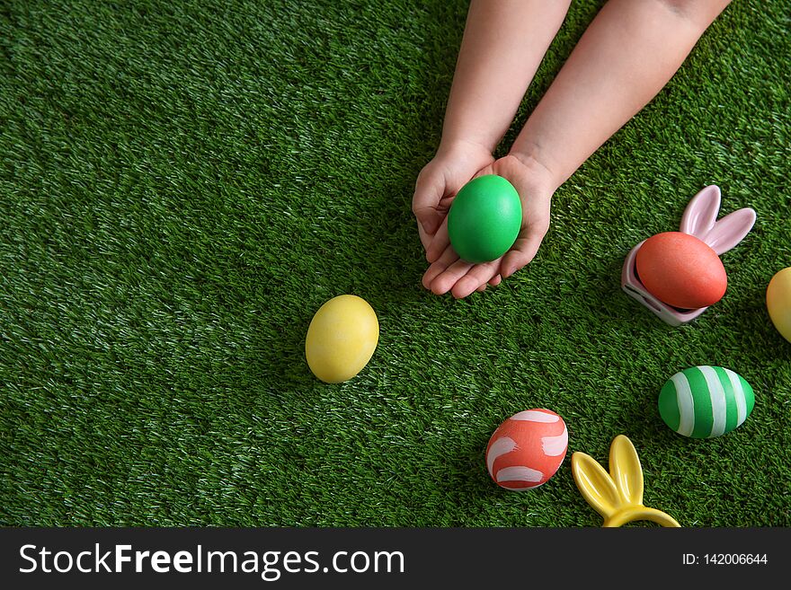 Little child holding painted Easter egg on green grass, top view.