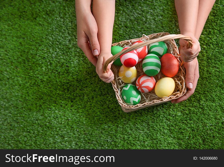 Mother and her little child with basket of painted Easter eggs on green grass, top view. Space for text