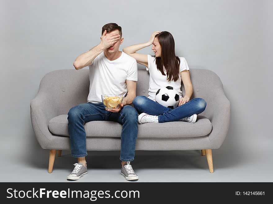 Displeased couple women men football fans cheer up support favorite team with soccer ball, holding bowl of chips, covering face with palm isolated on grey background. Sport family lifestyle concept. Displeased couple women men football fans cheer up support favorite team with soccer ball, holding bowl of chips, covering face with palm isolated on grey background. Sport family lifestyle concept