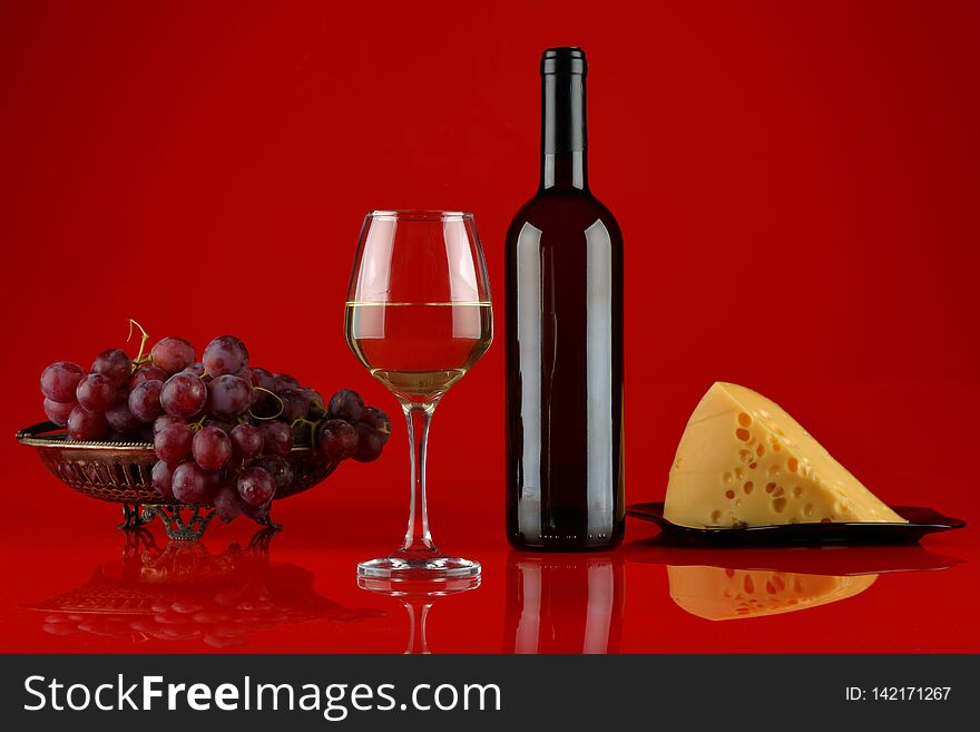 Still life with a glass and a bottle of white wine, a vase of grapes and cheese on a red, glossy background. Still life with a glass and a bottle of white wine, a vase of grapes and cheese on a red, glossy background