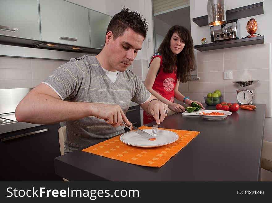 Hungry guy eating a slice of carrot while girl cooks.