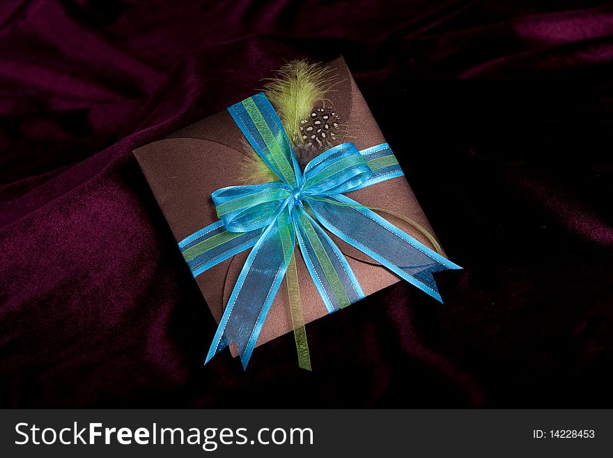 A brown gift folder with silk blue bow and guinea fowl feather on a maroon background. A brown gift folder with silk blue bow and guinea fowl feather on a maroon background