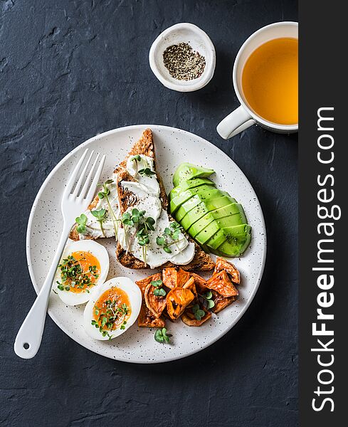Cream cheese toast, avocado, boiled egg, baked sweet potatoes and tea - delicious healthy breakfast snack on a dark background