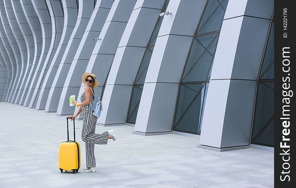 Young stylish Caucasian woman at the airport with a suitcase and a straw hat.