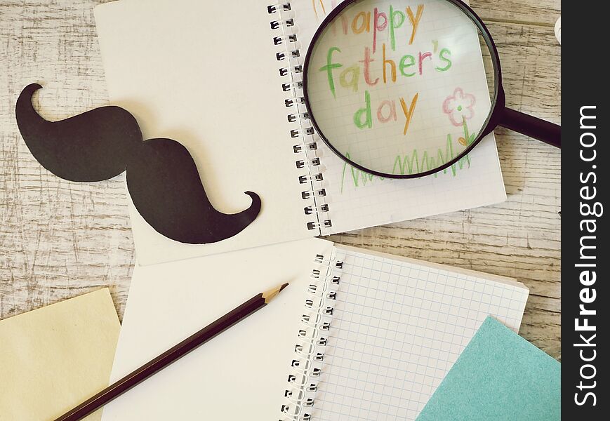 Father`s tools, black paper mustache, magnifying glass, pencils and a greeting inscription in a notebook on a light wooden table, top view, father`s day greeting concept, vintage style