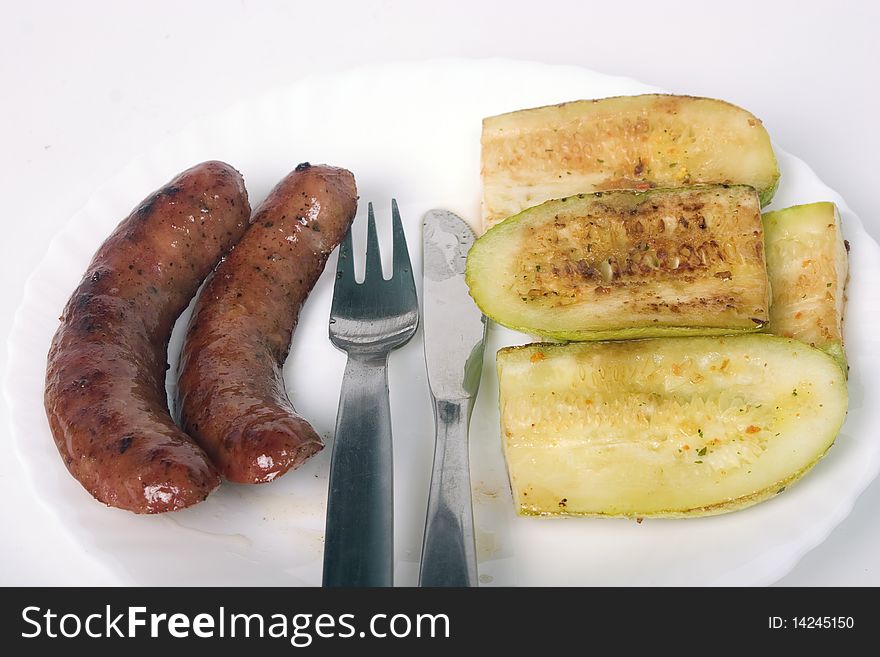 Two grilled sausages and zucchini on white plate with fork and knife between them. Two grilled sausages and zucchini on white plate with fork and knife between them