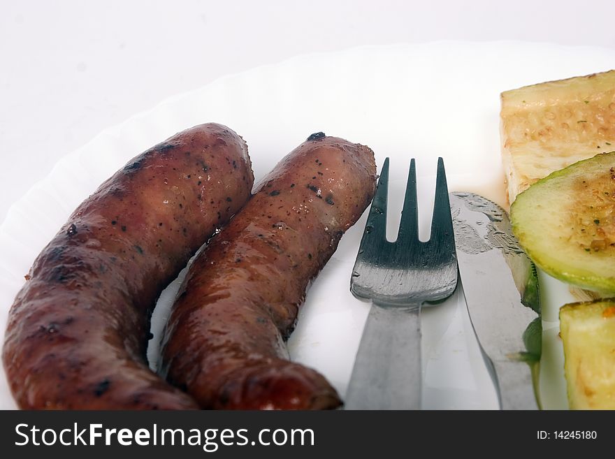 Two grilled sausages and zucchini on white plate with fork and knife between them. Two grilled sausages and zucchini on white plate with fork and knife between them