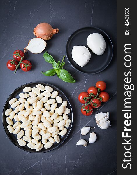 Gnocchi and mozarella on a black plate  and on a dark kitchen counter. Dark background. Ingredients for a baked gnocchi recipe. Tomato vines, garlic, onions, basil. Traditional Italian ingredients. Gnocchi and mozarella on a black plate  and on a dark kitchen counter. Dark background. Ingredients for a baked gnocchi recipe. Tomato vines, garlic, onions, basil. Traditional Italian ingredients.