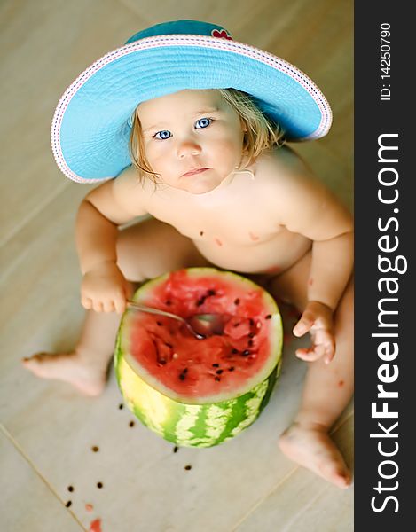 Cute baby-girl sits on a floor in a hat and eats a water-melon with a spoon. Cute baby-girl sits on a floor in a hat and eats a water-melon with a spoon