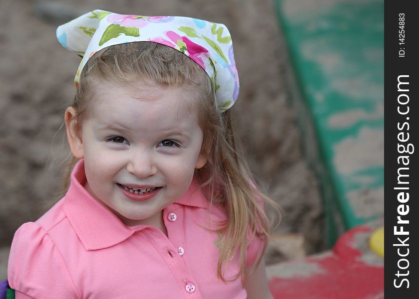 A Cute Girl Smiling in Sandbox with a Scarf on Head dressed in pink.