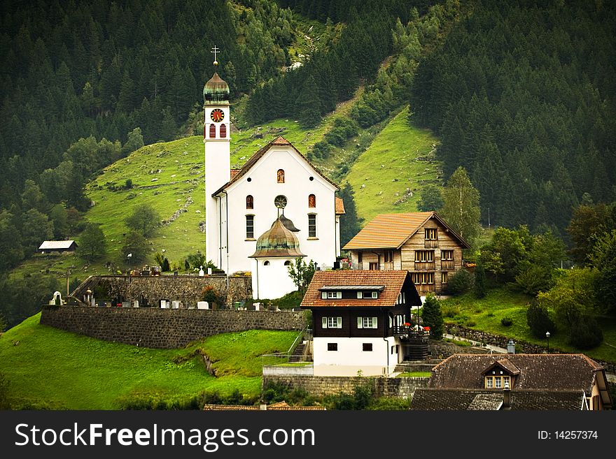 A church and several chalets surrounded by a green meadow and pine trees in the Swiss alps. A church and several chalets surrounded by a green meadow and pine trees in the Swiss alps