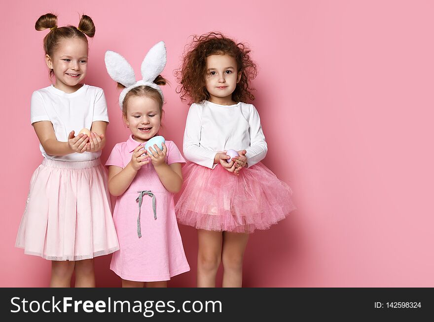 Three smiling dressed up for the holiday kid girls hold painted eggs on Easter day. Happy Easter celebration on pink background with free text space