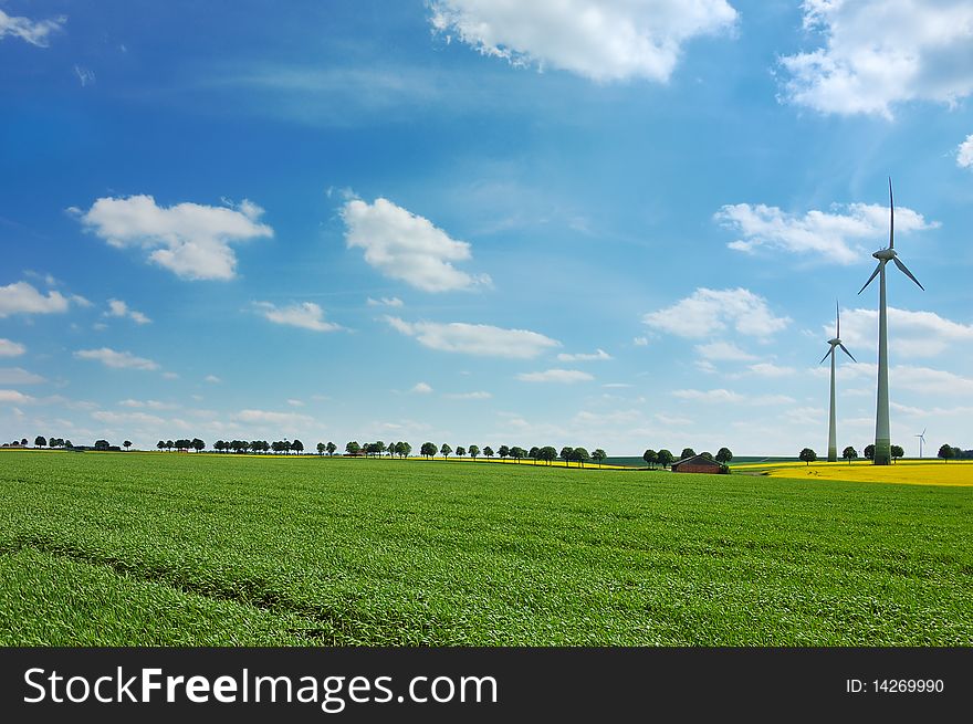 Wind turbines among rapeseed field and green meadows against a cloudy blue sky. Wind turbines among rapeseed field and green meadows against a cloudy blue sky