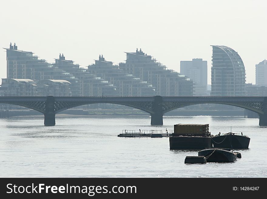 Futuristic buildings on the Thames river bank through some mist with bridge and barges in the foreground.