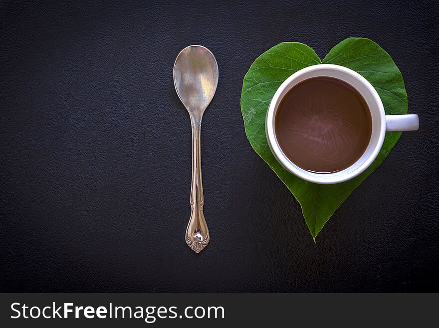 Top view image of a metallic spoon, a heart-shaped green morning glory leaf and a cup of coffee on black leather texture background. Top view image of a metallic spoon, a heart-shaped green morning glory leaf and a cup of coffee on black leather texture background.