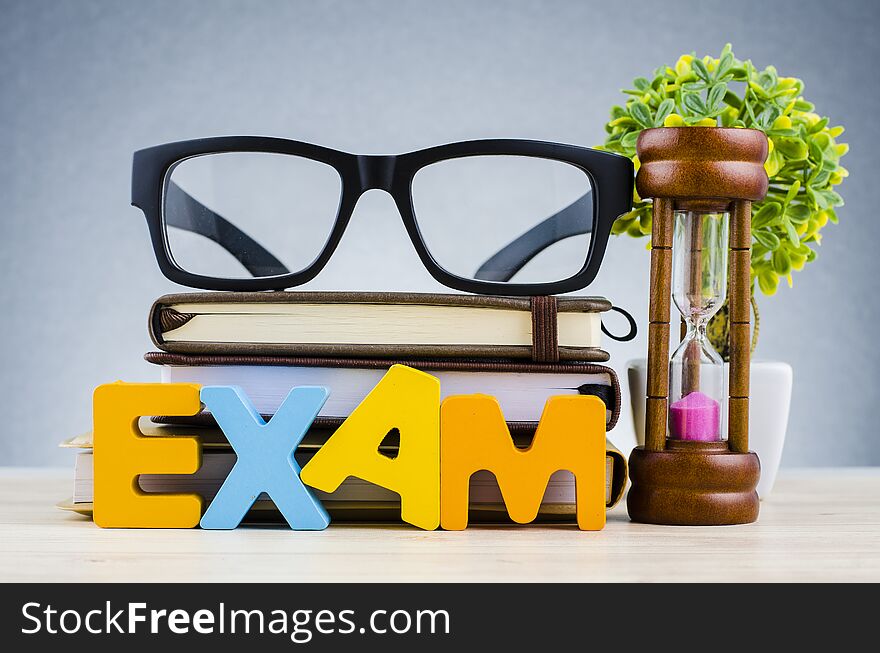 Education and time management concept, word EXAM over hourglass, and spectacles on stacking book