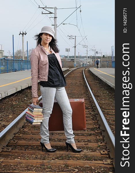 The young beautiful girl at station with a suitcase in a hand, in a light hat and a raincoat