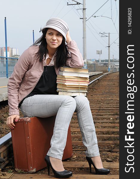 The young beautiful girl at station with a suitcase in a hand, in a light hat and a raincoat