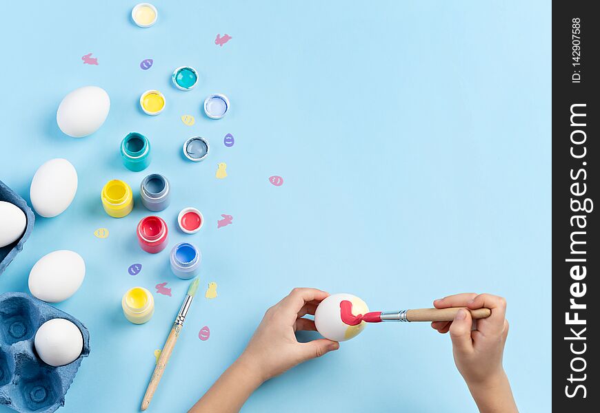 Close up image of kids hands painting an egg. Bottles of various paint, white eggs, bunny and chicken shaped sequins, brushes are around. Top view, space for text. Close up image of kids hands painting an egg. Bottles of various paint, white eggs, bunny and chicken shaped sequins, brushes are around. Top view, space for text.