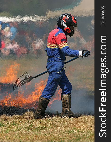 Man in a multi colored boiler suit, wearing a crash helmet, carrying a rake and trying to put out a fire on the grass. Man in a multi colored boiler suit, wearing a crash helmet, carrying a rake and trying to put out a fire on the grass.