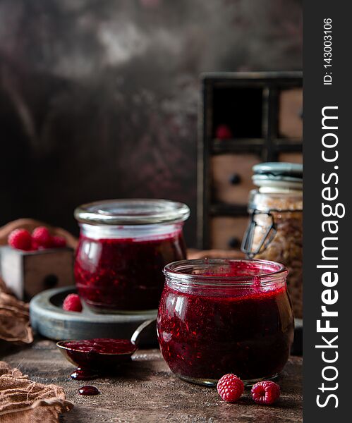 Two glass jars with homemade dark red jam or jelly standing on brown concrete table with raspberries, wooden board, chest of drawers, brown towel and jar with brown sugar. Preservation. Raspberry jam.