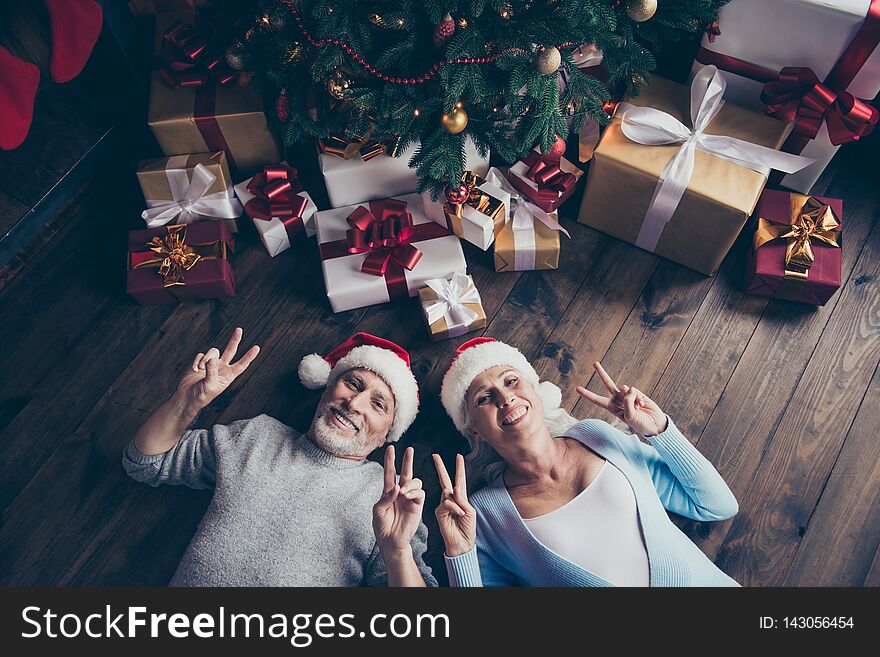 Top above high angle view of two adorable cheerful dreamy funny positive grey-haired spouses lying on floor near decorated fir tree among many bow ribbon boxes showing double v-sign symbol.
