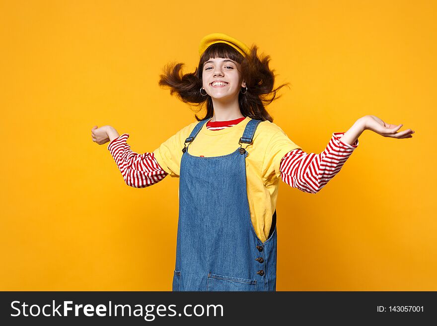 Cheerful girl teenager in french beret denim sundress spreading hands with fluttering hair isolated on yellow wall