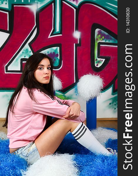 Lovely cheerleader wearing pink sweater and jeans shorts relaxing on the pom-poms. Graffiti background.