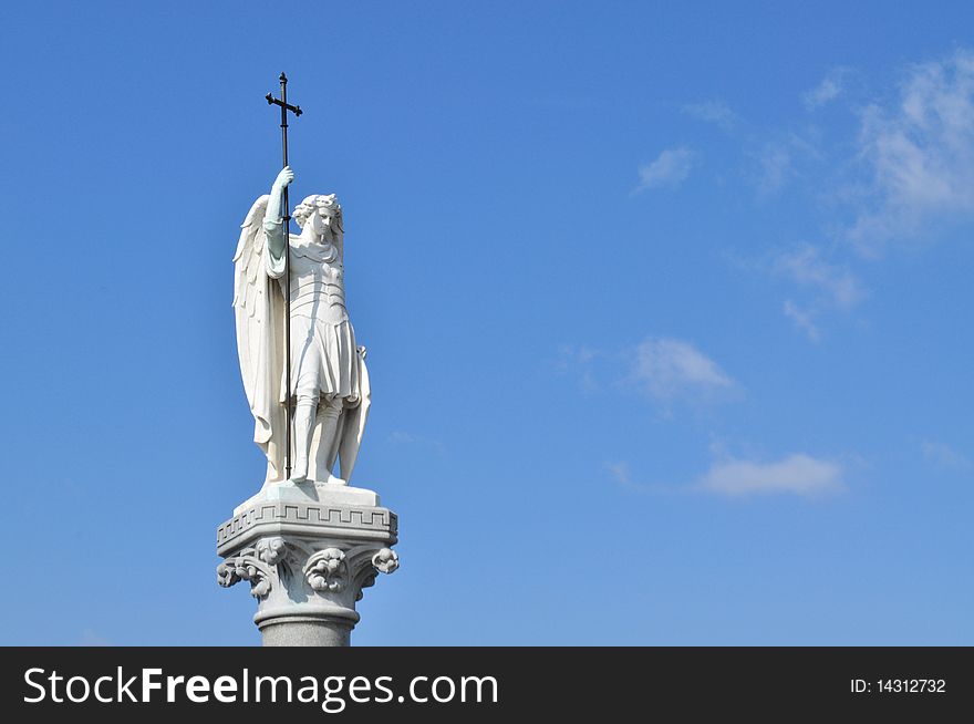 Statue of a winged holy man holding a cross
against a blue sky. Statue of a winged holy man holding a cross
against a blue sky
