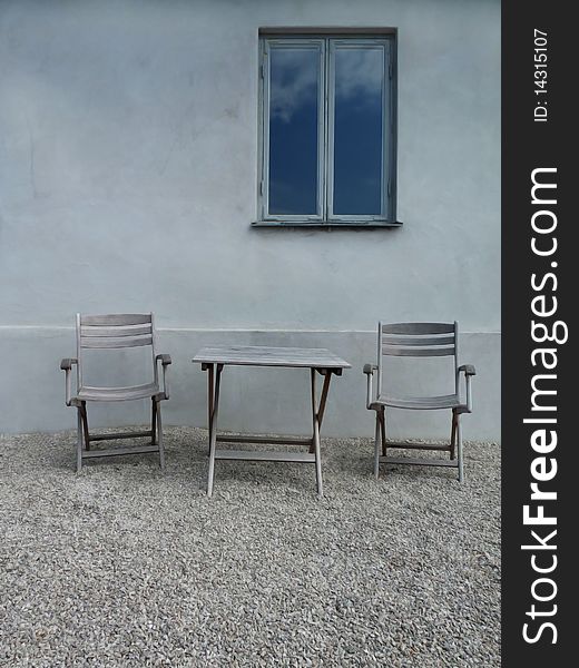 Two chairs and a table on gravel towards plaster wall with reflecting window. Two chairs and a table on gravel towards plaster wall with reflecting window.