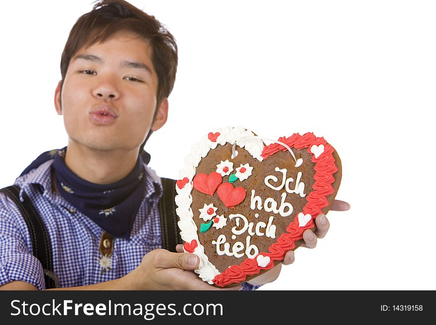 Chinese man, dressed with Bavarian Lederhose holds an Oktoberfest gingerbread heart in camera and gives a kiss. Isolated on white. Chinese man, dressed with Bavarian Lederhose holds an Oktoberfest gingerbread heart in camera and gives a kiss. Isolated on white.