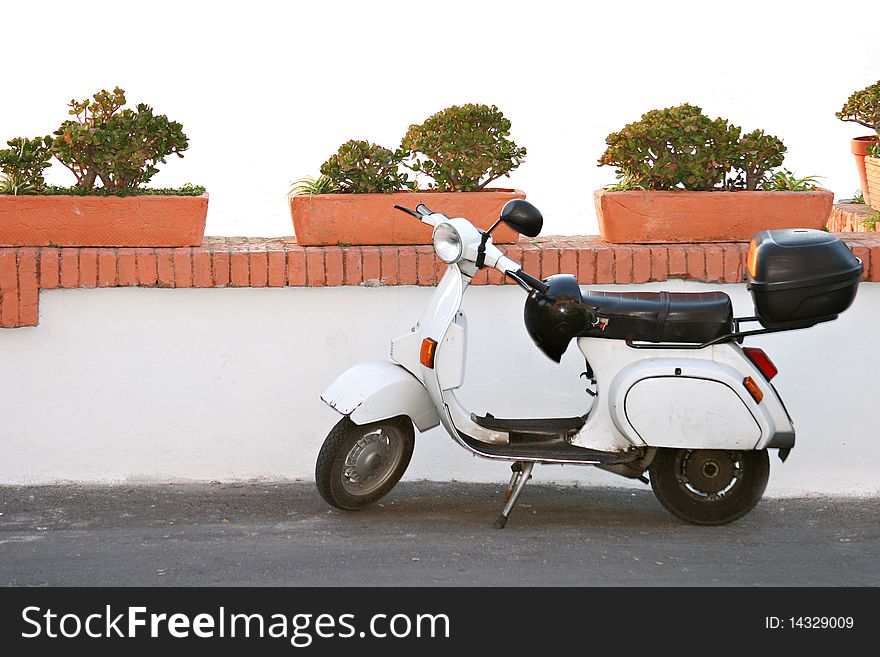 A white and black motorbike is parked alongside a white wall with red brick trim and red terracotta planter boxes. Italy. A white and black motorbike is parked alongside a white wall with red brick trim and red terracotta planter boxes. Italy.