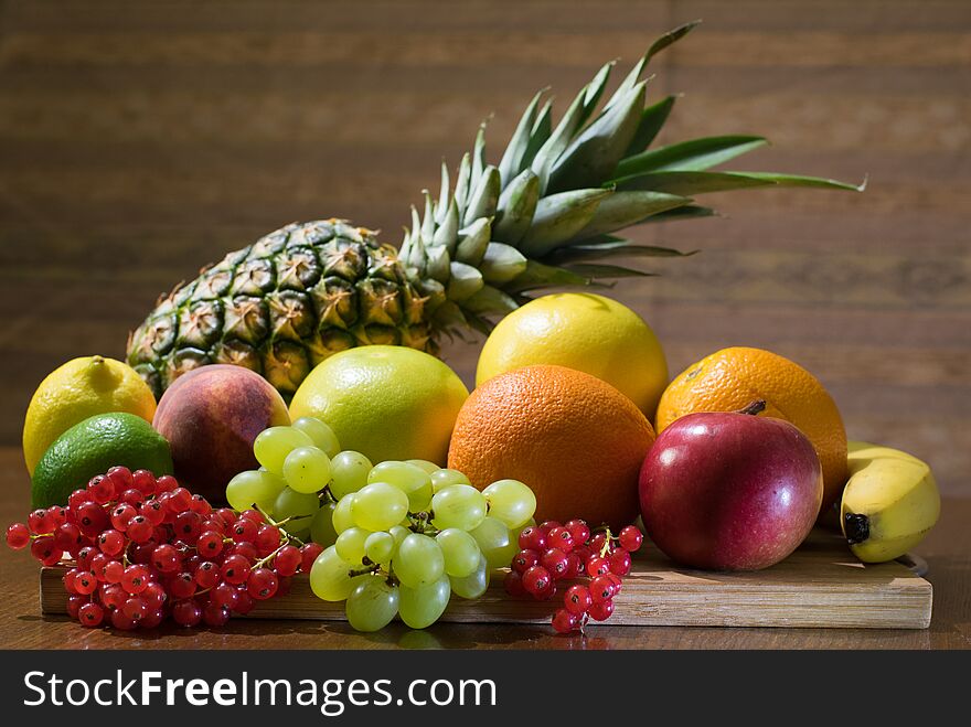 Different kinds of fruits, pineapple, grapes, currant, apple, orange grapefruit, lime, lemon, peach, banana, on the wooden board at the table with brown background. Different kinds of fruits, pineapple, grapes, currant, apple, orange grapefruit, lime, lemon, peach, banana, on the wooden board at the table with brown background.