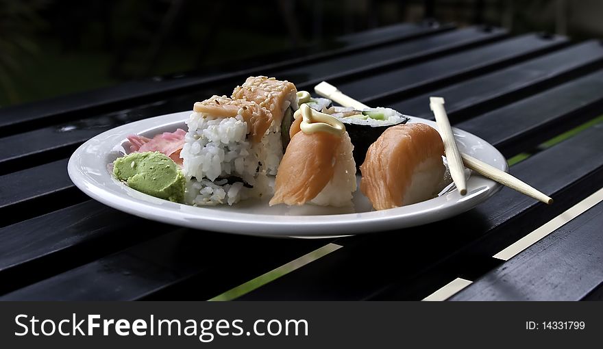 A plate of sushi on a white plate lying on a black wood table with eating utensils