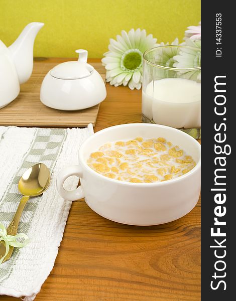 On the wooden table is breakfast cereal with milk, a glass of milk, a spoon, napkin, tea and flowers