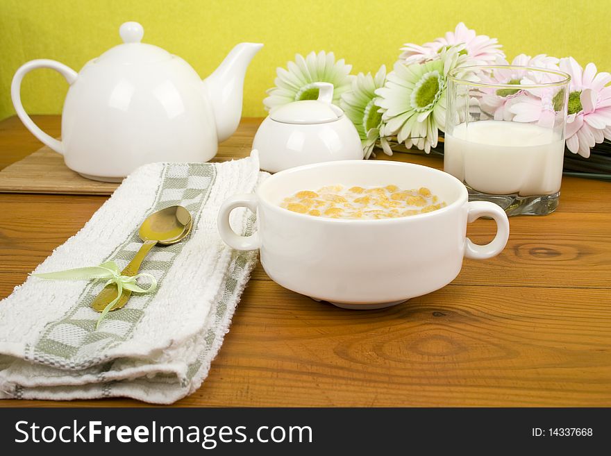 On the wooden table is breakfast cereal with milk, a glass of milk, a spoon, napkin, tea and flowers