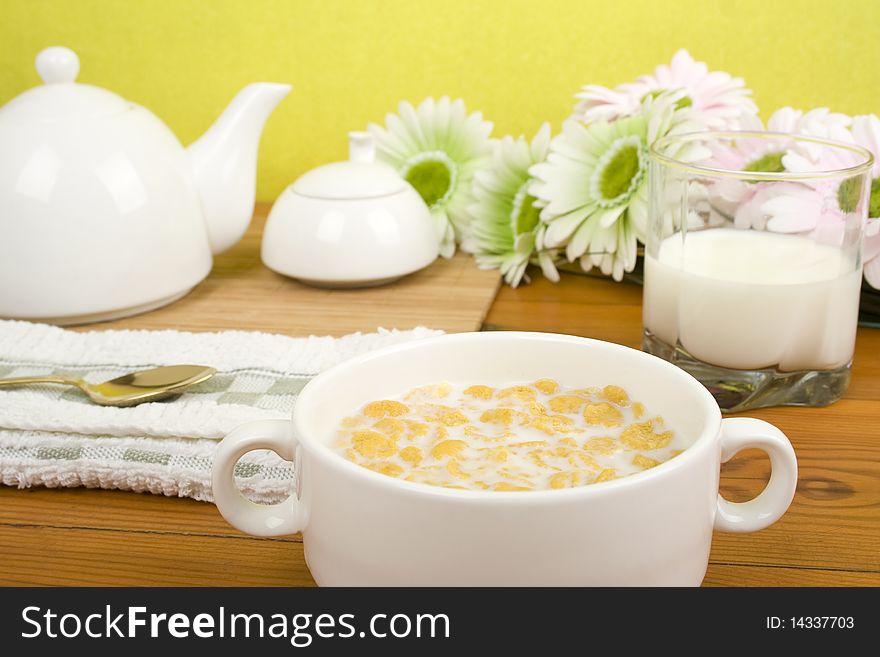 On the wooden table is breakfast cereal with milk, a glass of milk, a spoon, napkin, tea and flowers